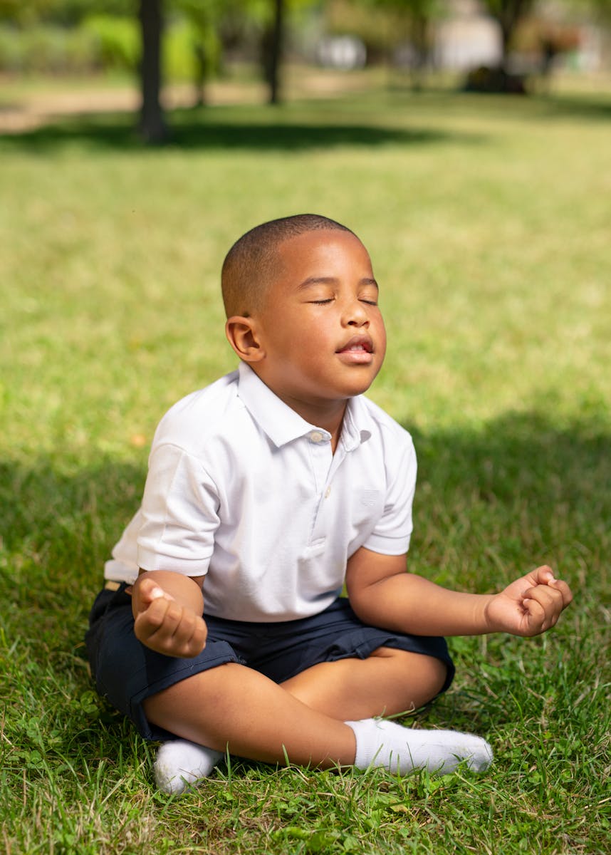 A young boy meditates peacefully on a grassy field during a sunny day, eyes closed and relaxed.