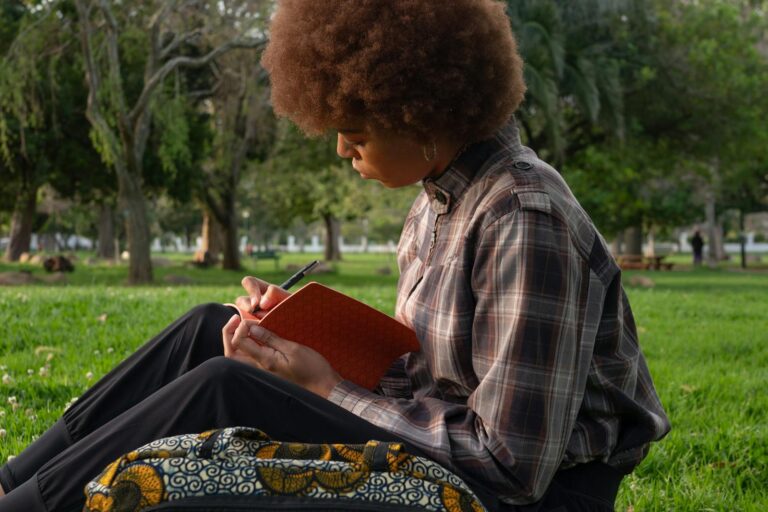A young woman with curly hair writing in a notebook outdoors, embodying leisure and creativity.